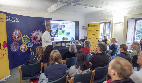 A low resolution image of Professor Ian Thompson stood in front of roller banners and a screen with a PowerPoint presentation on it, presenting to a small room of people.